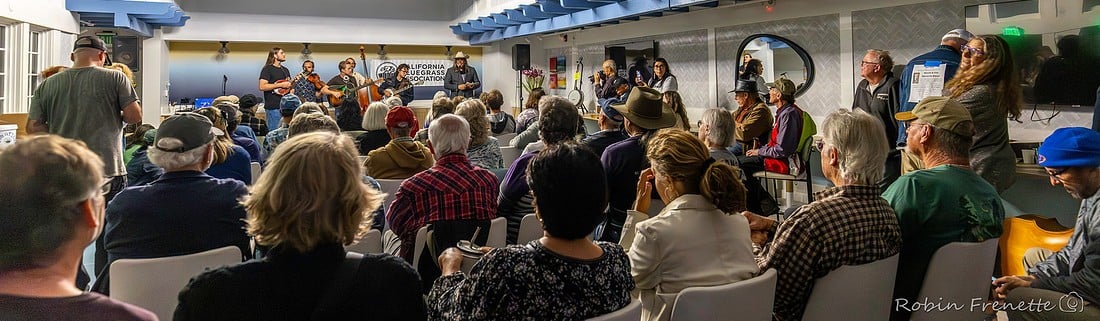 A crowd sits in a brightly lit room watching a live band perform on a small stage, with various audience members focused on the musicians.
