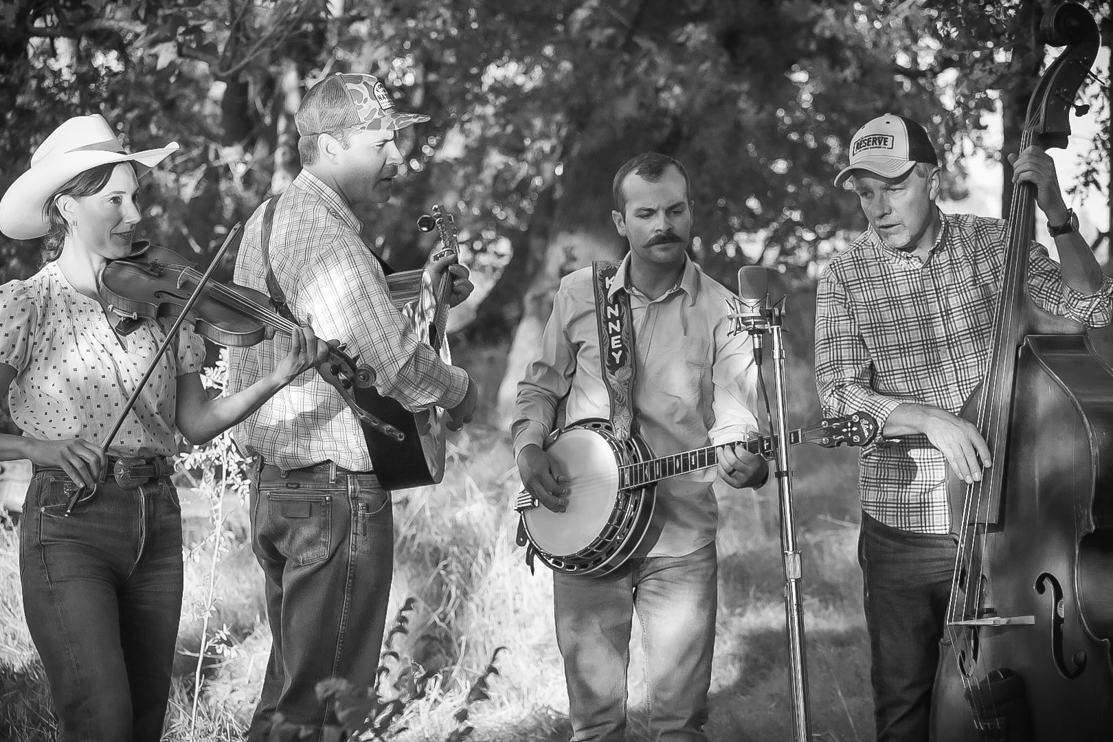 Four musicians play fiddle, guitar, banjo, and upright bass outdoors, standing closely together around a microphone.