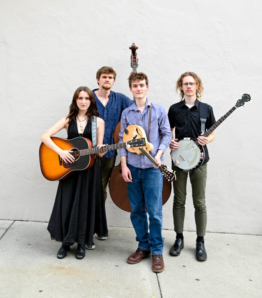 Four musicians stand against a plain wall, holding a guitar, upright bass, mandolin, and banjo.