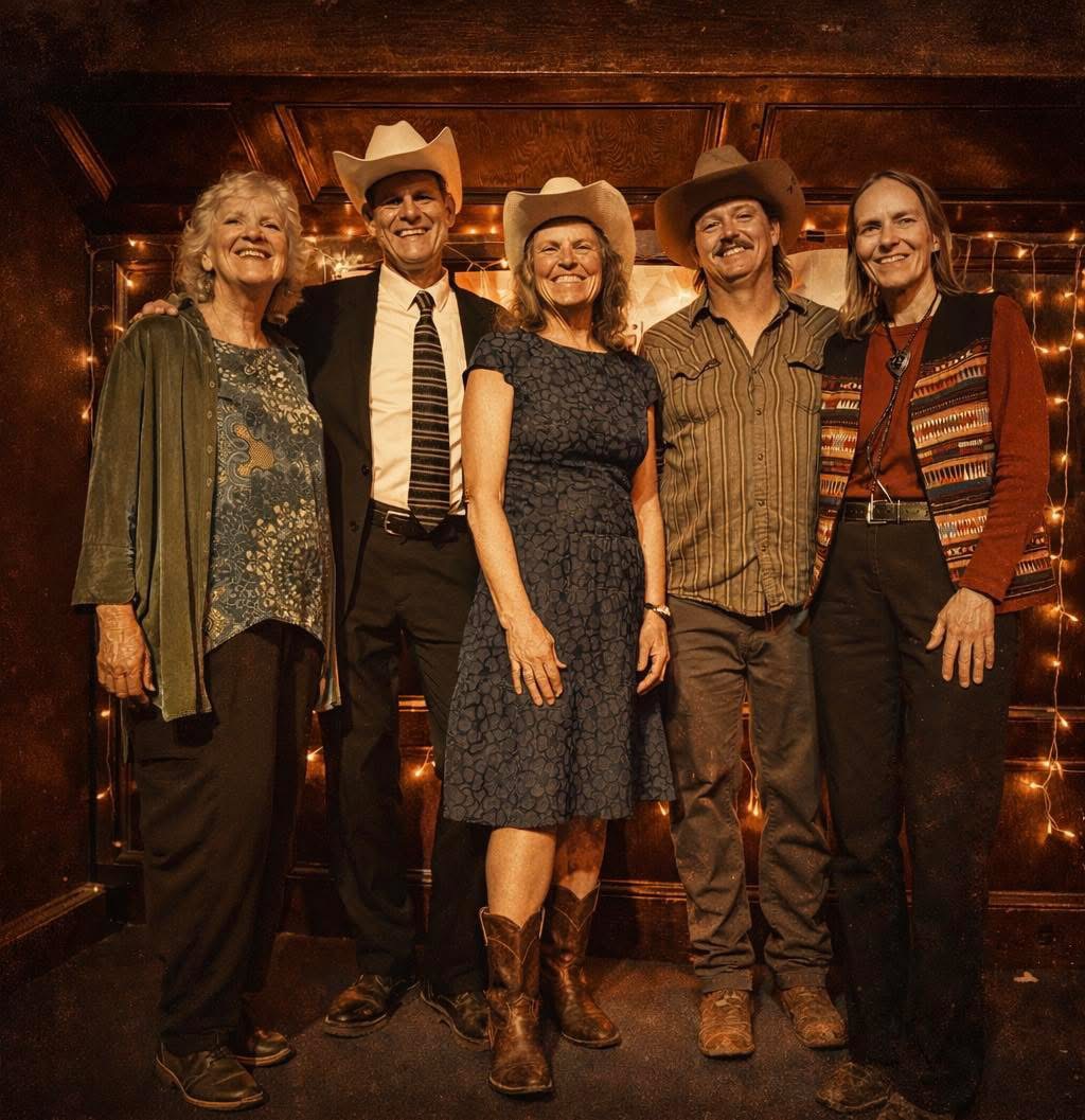 Five adults stand together indoors, dressed in semi-formal and country-style clothing, including cowboy hats and boots, with string lights in the background.