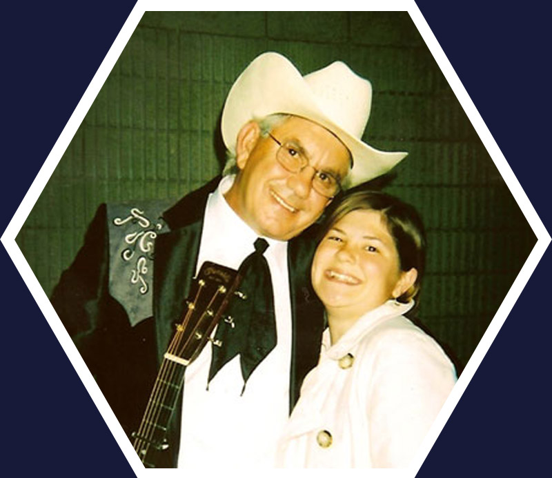 An older man in a cowboy hat and suit with a guitar stands smiling next to a young girl in a light-colored coat, both posing for the camera.