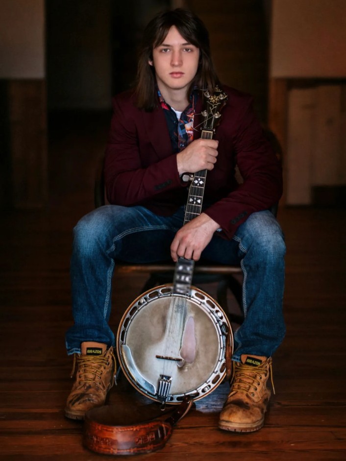 Ettore Buzzini Seated Banjo A young person with long hair sits on a chair, holding a banjo upright and resting one hand on its neck. They wear a burgundy jacket, jeans, and work boots.