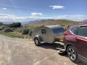 A red SUV is towing a small silver teardrop trailer parked on a gravel road, with shrubs, hills, and distant mountains under a partly cloudy sky.
