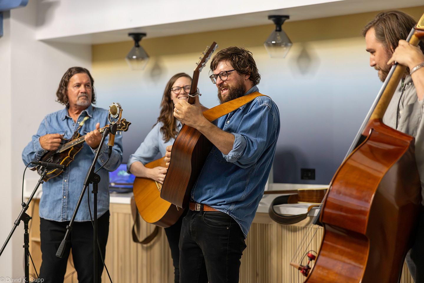 2025 South State 48 Four musicians perform indoors; one plays guitar and sings, another plays upright bass, another plays mandolin, and one plays violin. All are smiling and wearing casual attire.