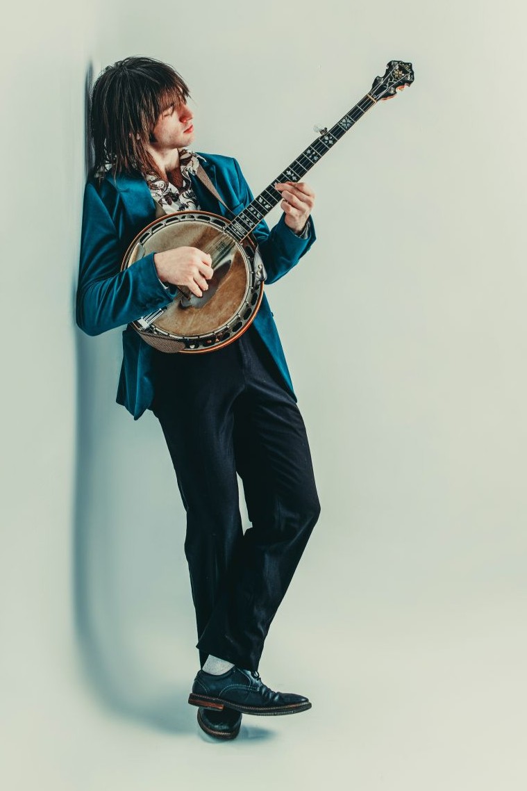 Ettore Rockabilly Lean 2026 cropped Person in a blue blazer and black pants leans against a wall, playing a banjo, with one foot resting on a small balance board.