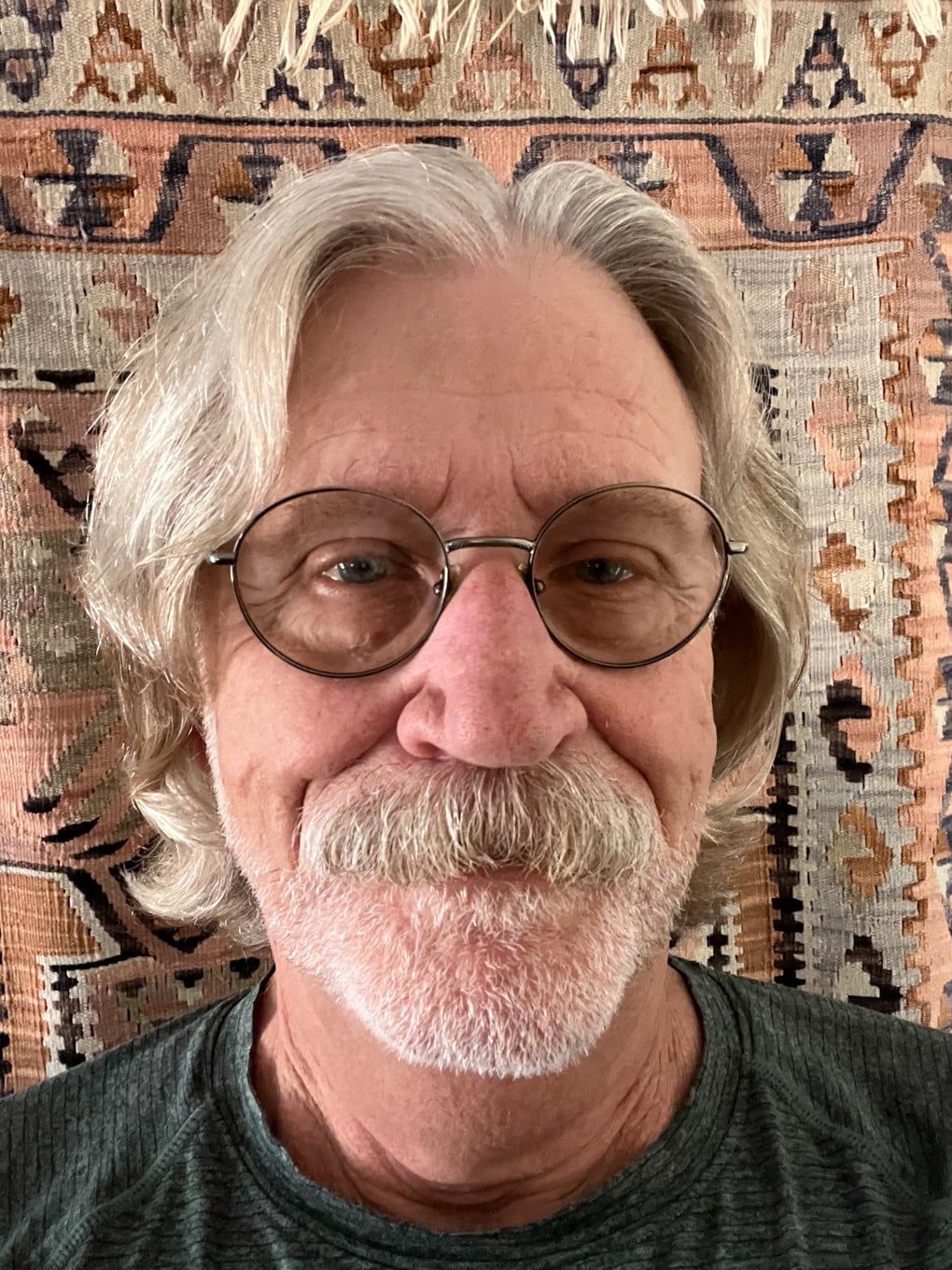 An older man with gray hair, glasses, and a mustache smiles slightly, standing in front of a patterned rug background.