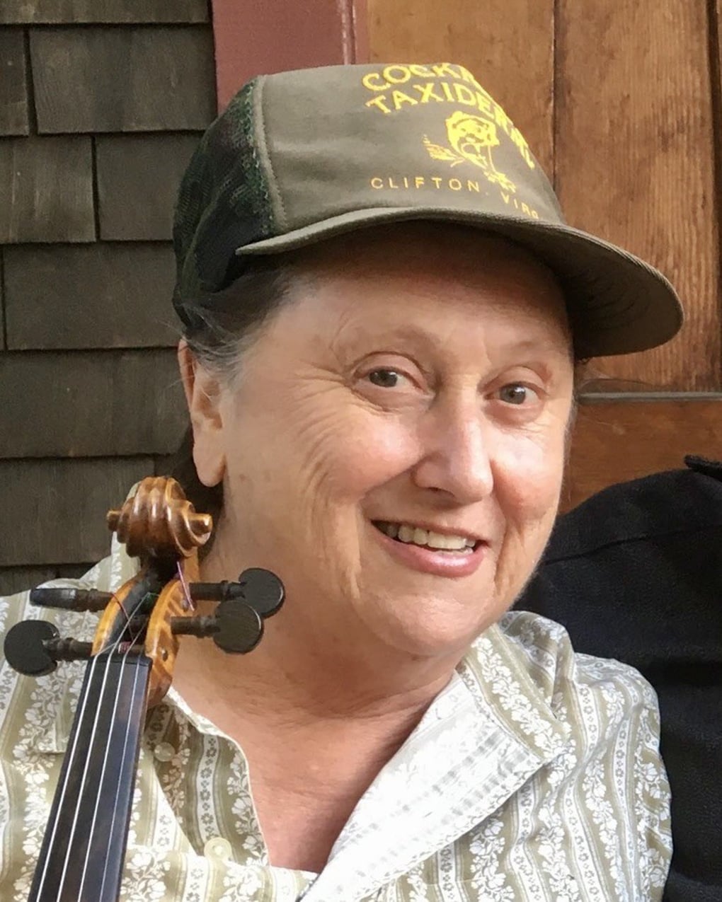Older woman wearing a green cap and patterned shirt, smiling while holding a string instrument, standing in front of a wooden background.