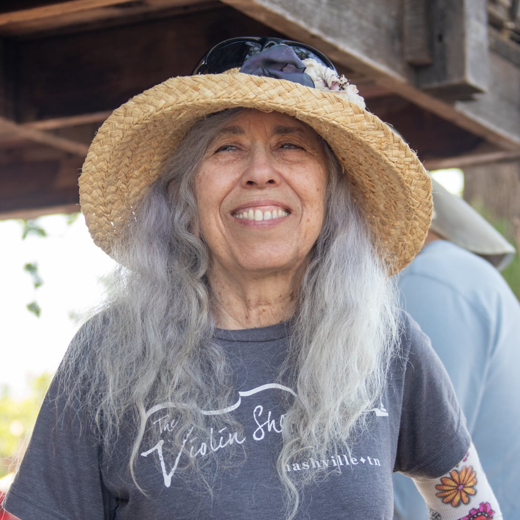 Older woman with long gray hair smiles, wearing a straw hat and a “The Violin Shop” t-shirt outdoors, with sunglasses resting on her hat.