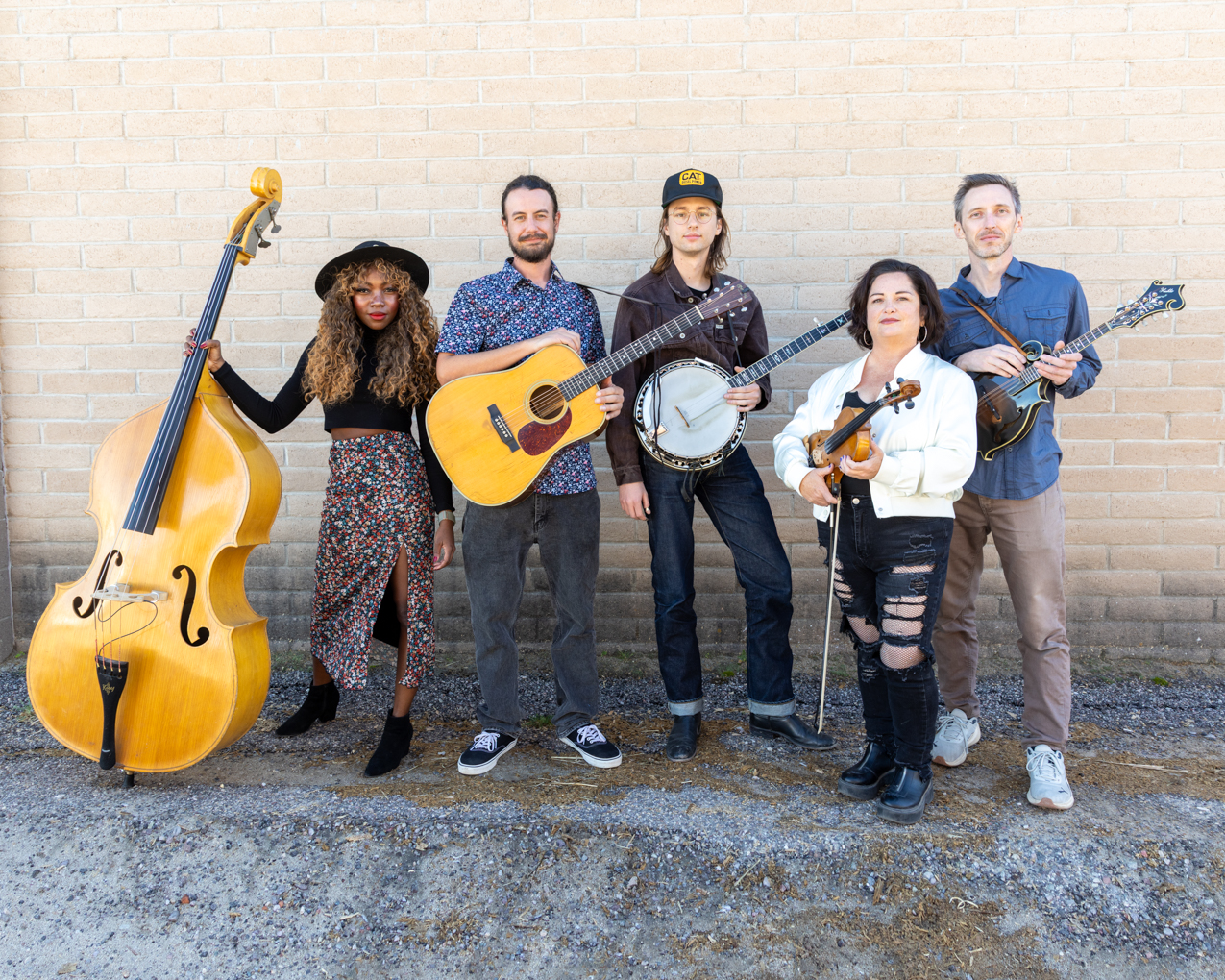 Five people stand in front of a brick wall holding musical instruments: double bass, acoustic guitar, banjo, violin, and mandolin.