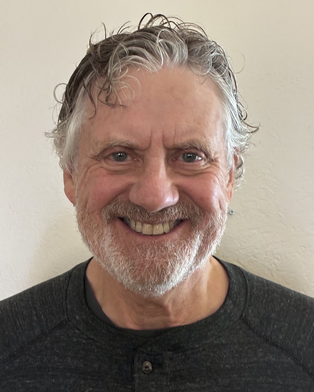 Smiling older man with gray hair and beard wearing a dark shirt stands against a plain light-colored wall.