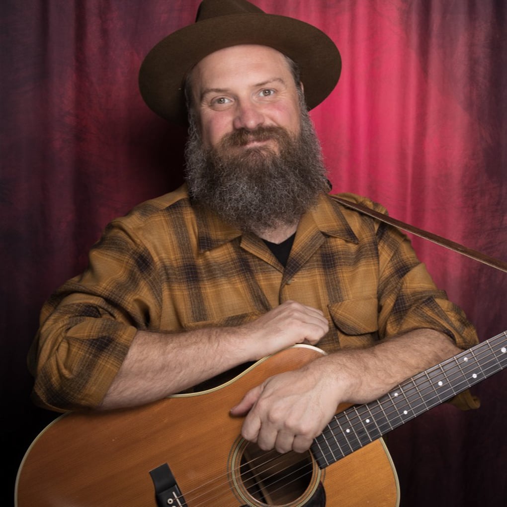 Man with a full beard wearing a brown hat and plaid shirt sits in front of a red backdrop, holding an acoustic guitar.
