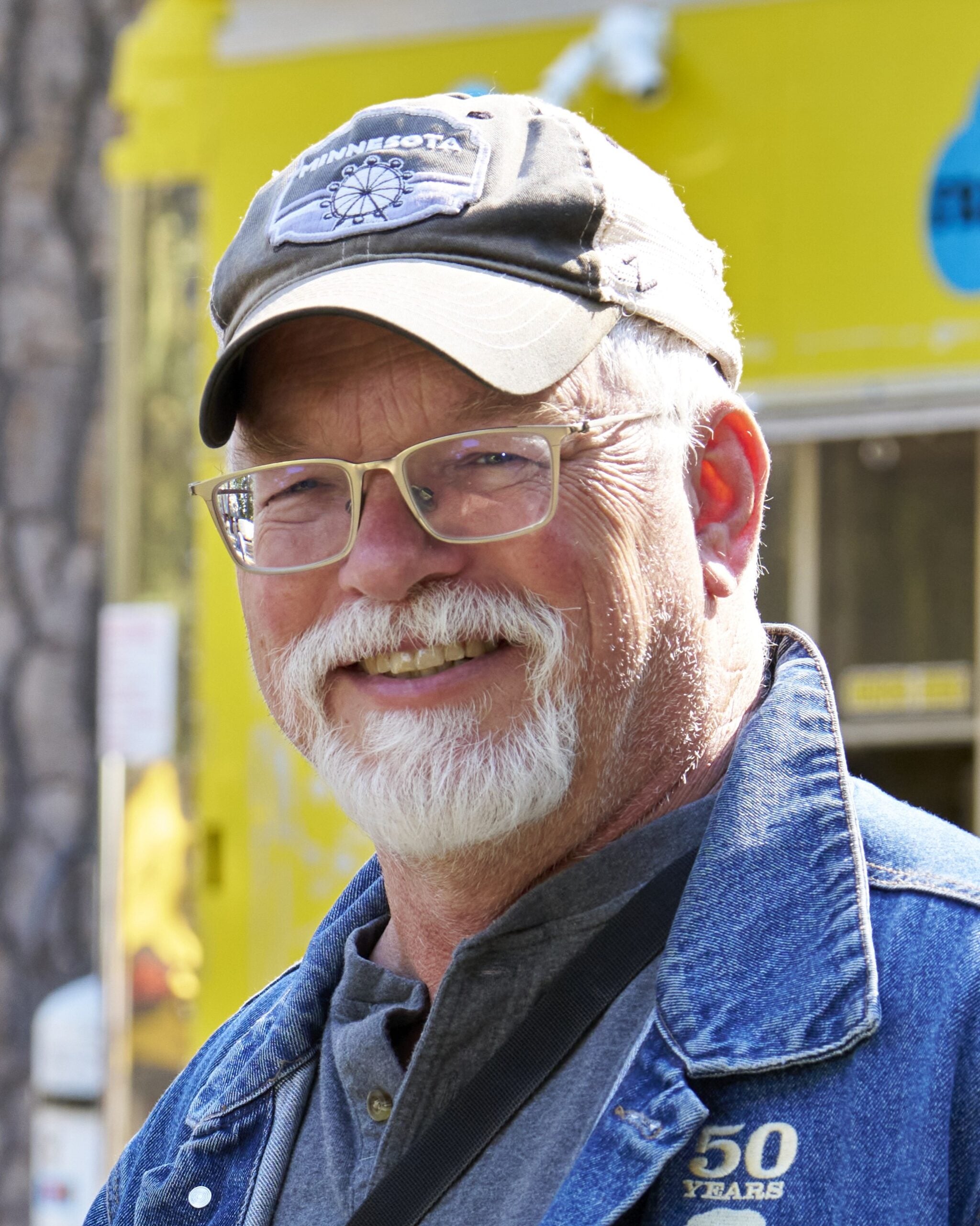 Older man with white hair, beard, and glasses stands in front of a vintage-style microphone, wearing a dark shirt. Trees and a building are visible outside the window behind him.