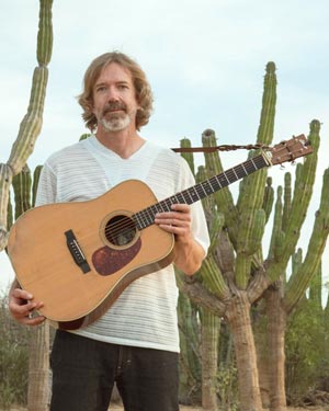 A person with a beard stands outdoors, holding an acoustic guitar in front of a background of tall cacti.