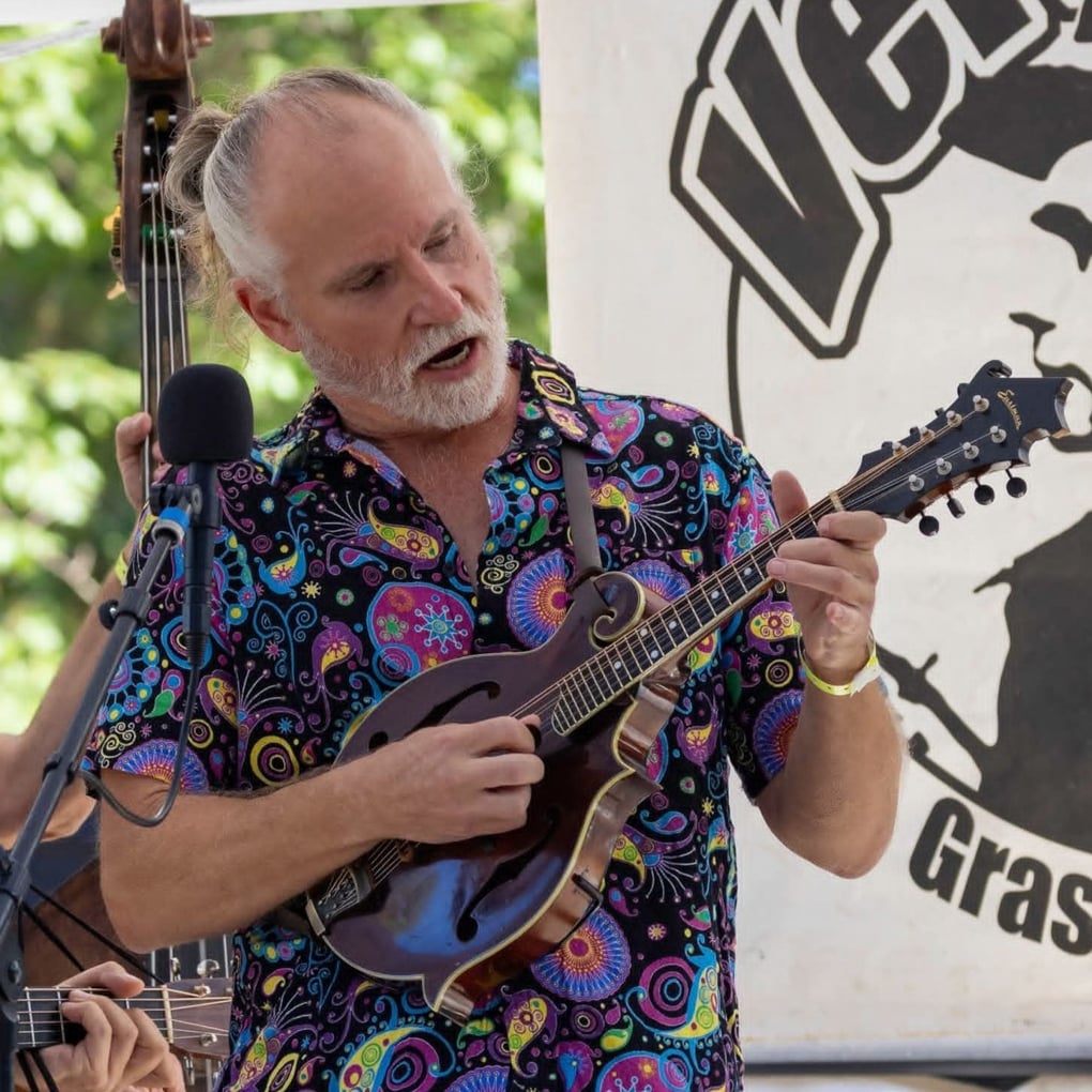 A man with gray hair and a colorful shirt plays a mandolin and sings into a microphone at an outdoor event.