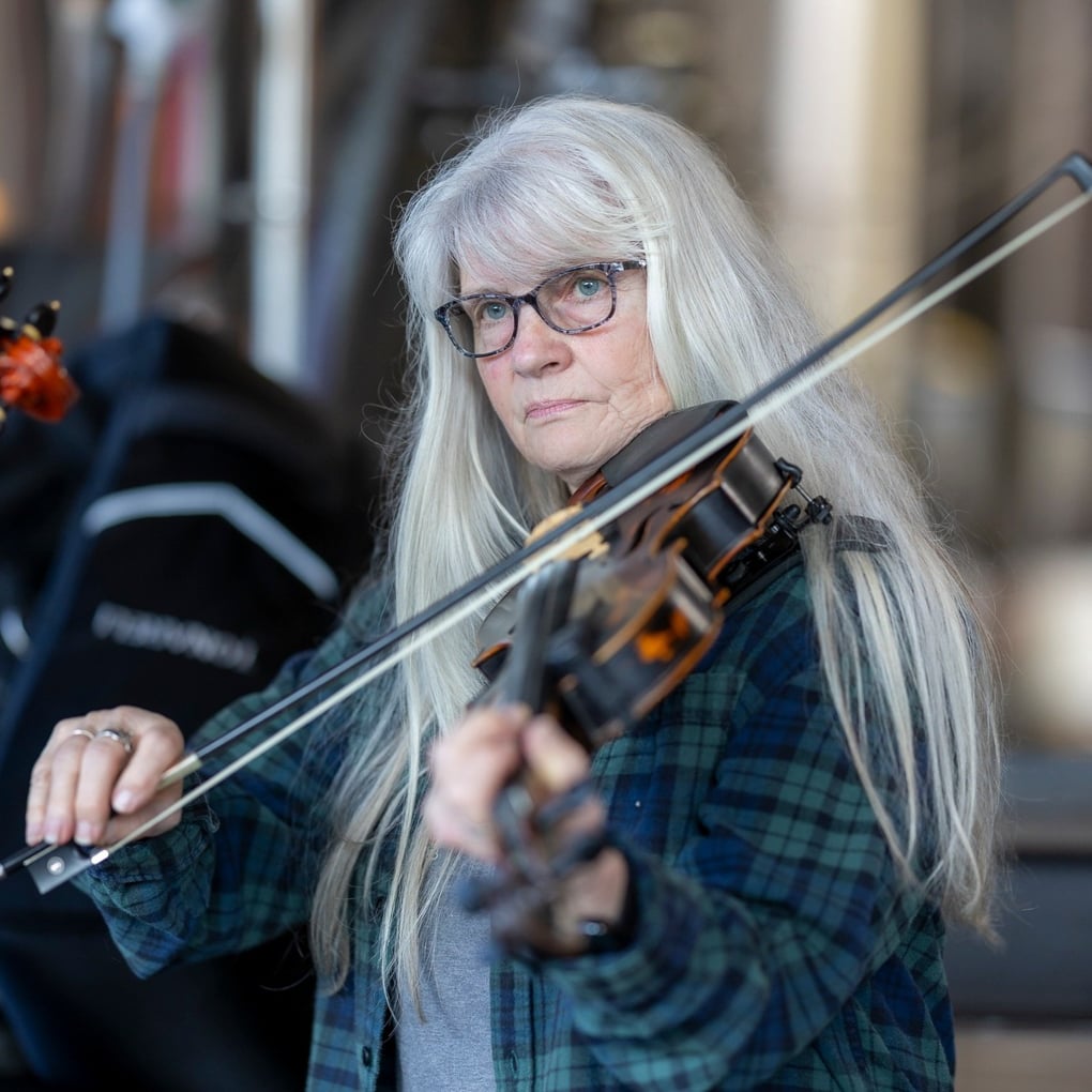 An older woman with long gray hair and glasses plays the violin, wearing a green plaid shirt and standing indoors.