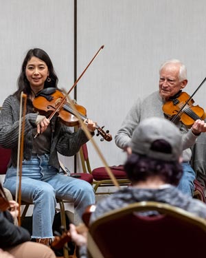Two people playing violins in a music group setting with an audience in front of them.