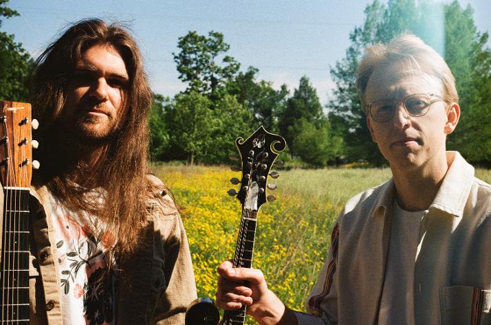 Two men stand in a field of yellow flowers holding stringed instruments, with trees and blue sky in the background.