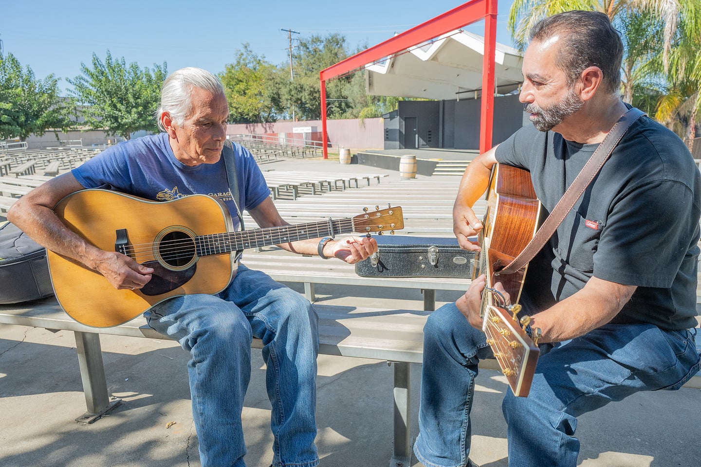 Two men sitting on outdoor benches playing acoustic guitars.